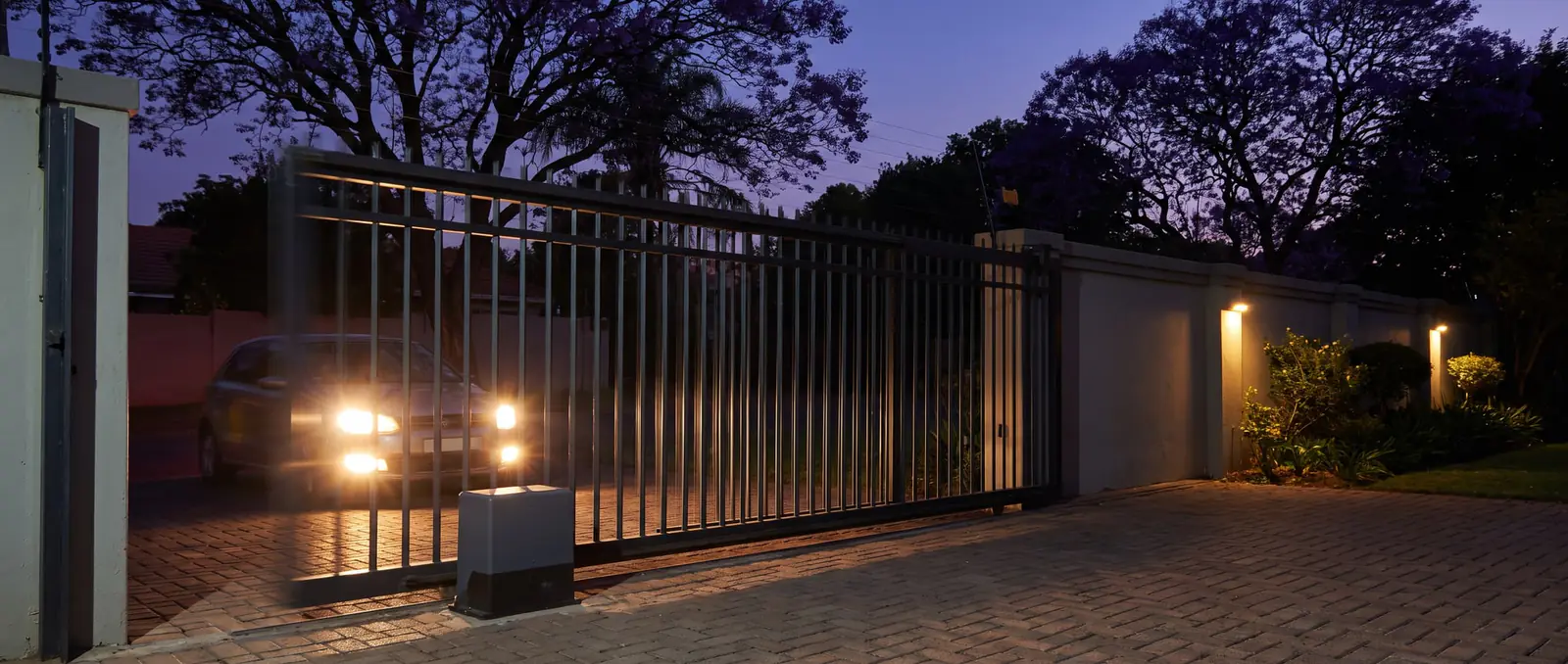 A black steel sliding gate opening across a driveway at dusk, a car's headlights waiting outside